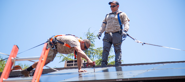 Two installers bolting solar panel to dark shingle roof.
